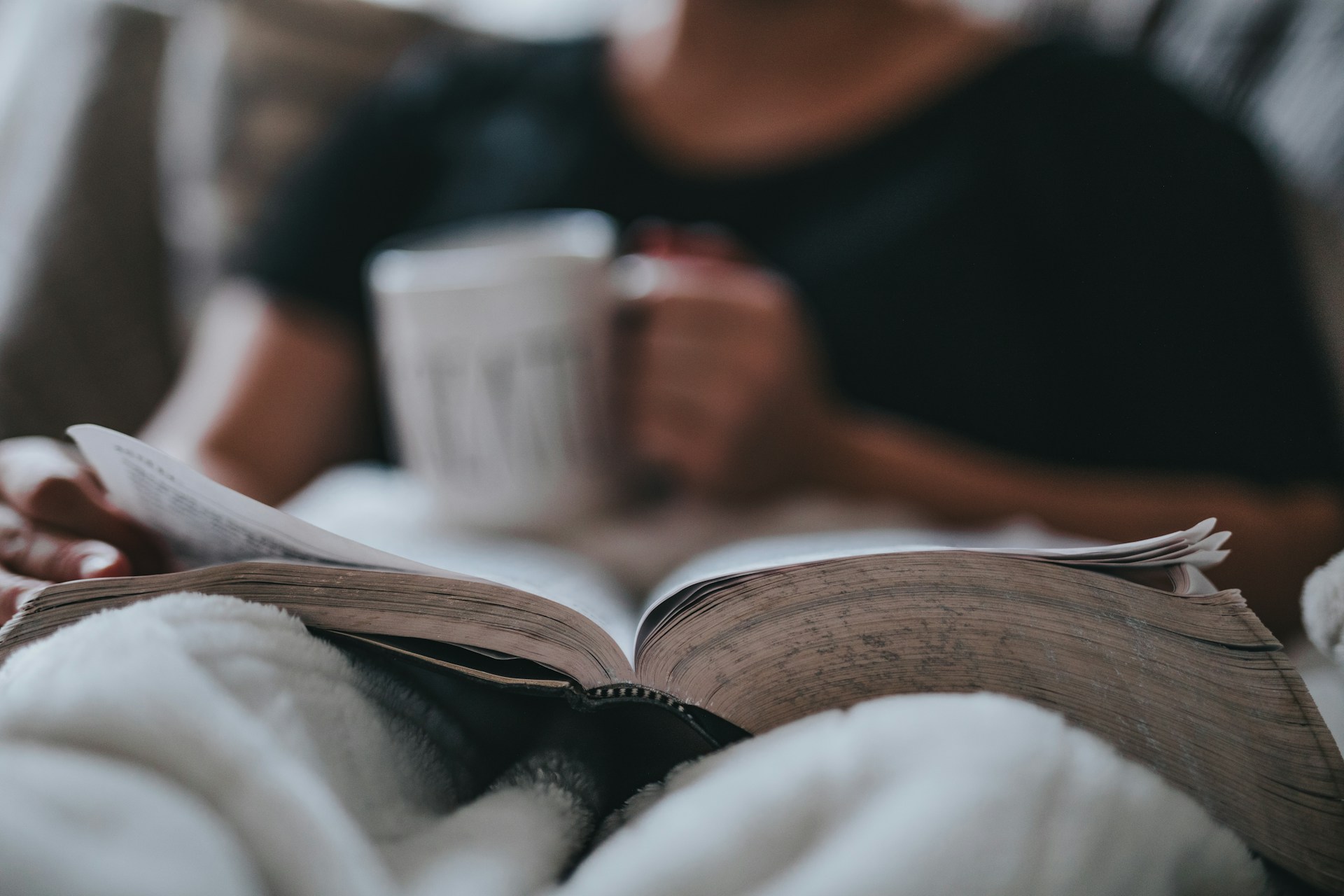 a person holding a coffee mug and reading a book on the sofa with a blanket enjoying some downtime